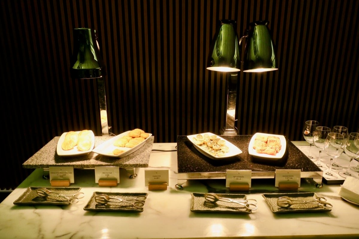 Hot food buffet at Cathay Pacific The Wing First Class Lounge in Hong Kong International Airport, featuring Chinese steamed egg cake, BBQ duck puff, spinach dumplings, assorted danish pastries, and wine glasses on a marble counter.