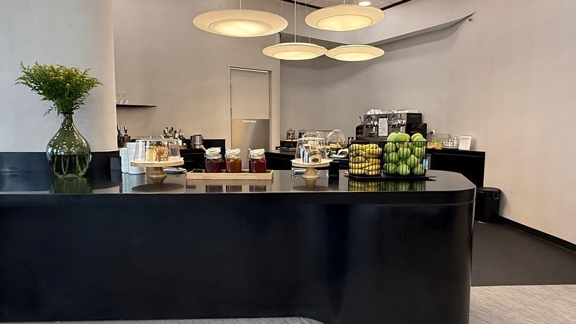 Food station at the Goldair x aia lounge at Athens Airport. The station has glossy black counters with a variety of food items including fresh fruit, condiments and pastries under glass covers. There is a coffee maker and alcoholic drinks in the back