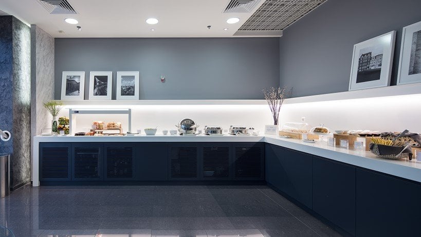 Modern buffet area in the goldair cip lounge at Athens Airport. It has a clean minimalist design with grey and navy blue colour scheme. The buffet counter has various food items and snacks, and is illuminated under a cabinet light.