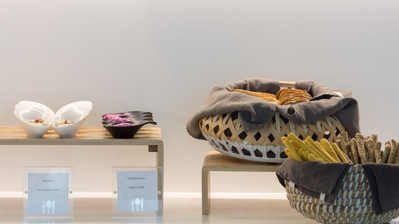 Close up view of the buffet section in the Goldair CIP lounge at Athens Airport. The setup includes a wooden bench showcasing various food items including vibrant beetroot salad or hummus, bread rolls, and crisp breadsticks in woven baskets.