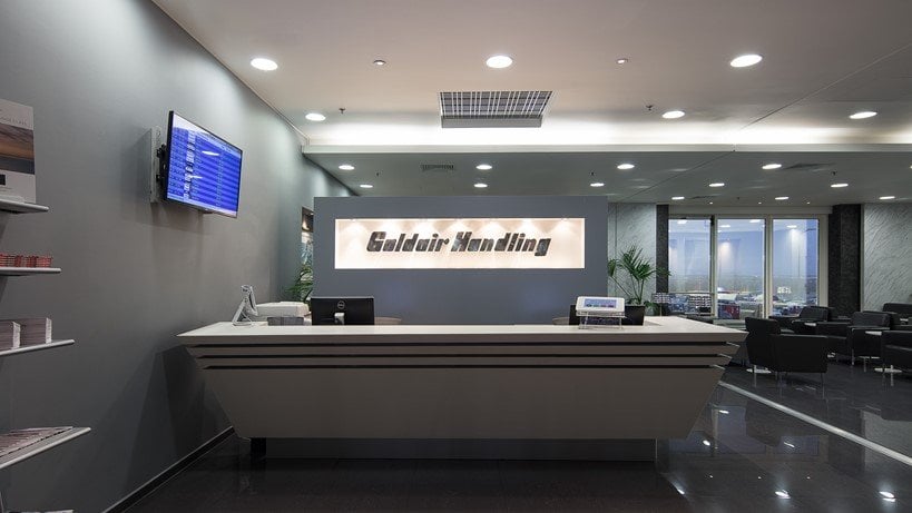 Reception desk at the Goldair Handling CIP Lounge at Athens Airport. The front desk is minimalist with illuminated signage against a grey backdrop. To the side is a digital flight information display, and a seating area with window views.
