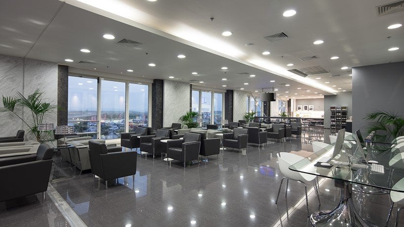 Spacious and modern seating area in the Goldair Handling CIP lounge at Athens Airport, featuring a wide array of grey armchairs arranged neatly around glass tables. Large windows along the room provide panoramic views of the countryside.