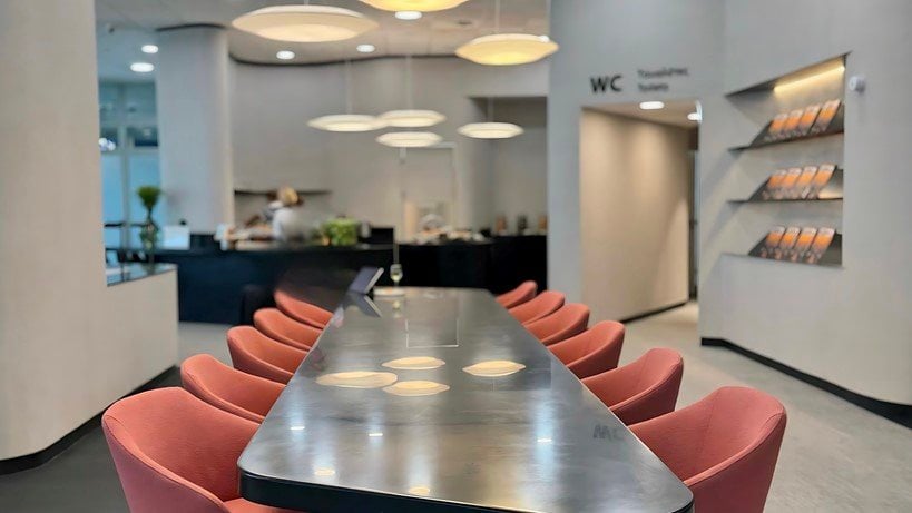 Dining area of the Goldair x aia lounge in Athens Airport. Featuring a modern and chic metallic table surrounded by salmon coloured chairs. The background shows a well lit buffet area with a variety of dishes on display. On the right wall are flyers.