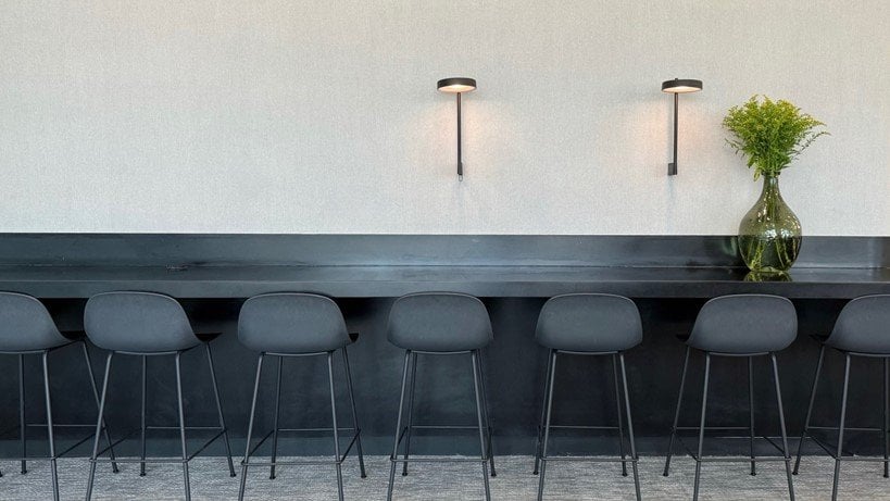 Modern and minimalistic seating area at the Goldair x aia lounge at the Athens Airport. This photo features a low long bar table and plenty of bar stools in a deep grey colour. There is decorative greenery in a glass plant holder on the right.