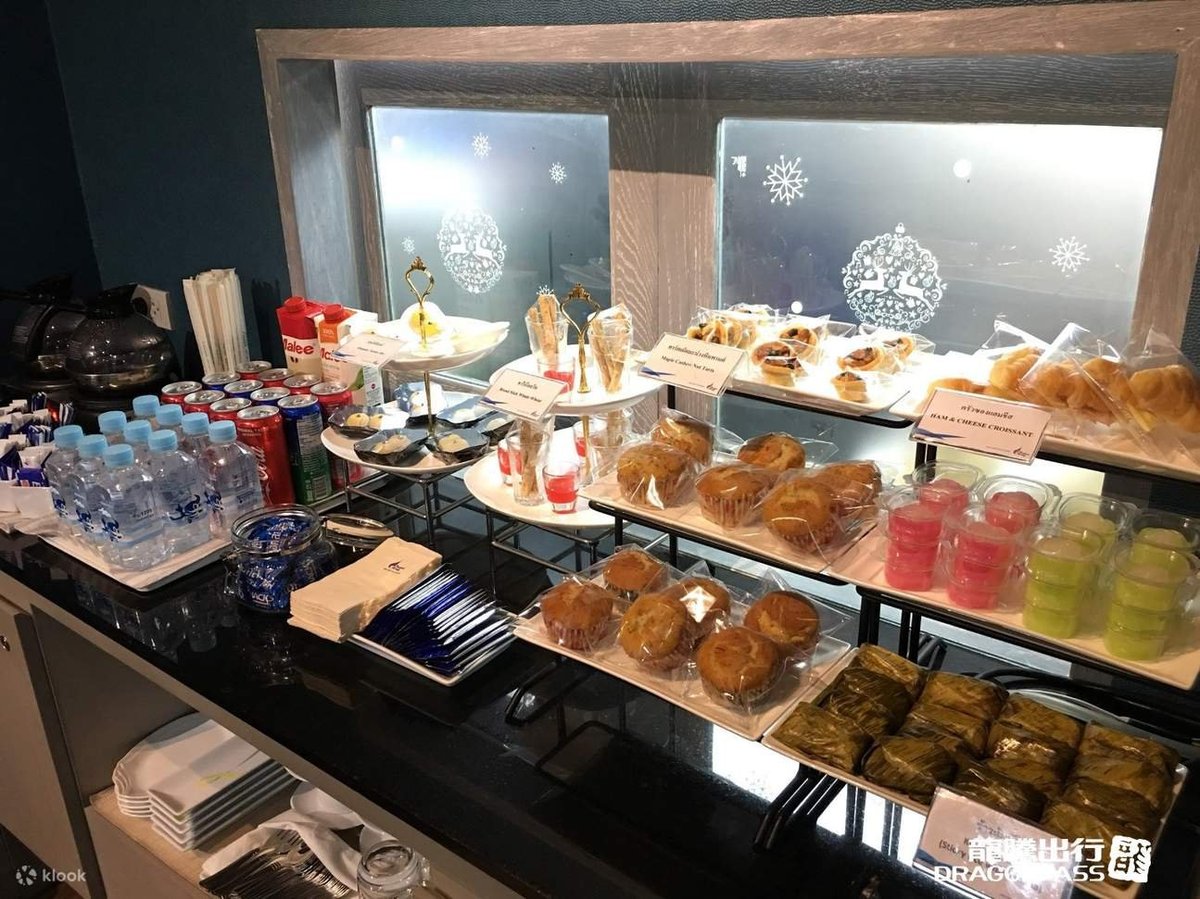 Buffet station at Chiang Rai Airport Coral Executive Lounge with an assortment of snacks including ham and cheese croissants, muffins, Thai desserts, bottled water, soft drinks, and coffee- self-serve refreshment area.