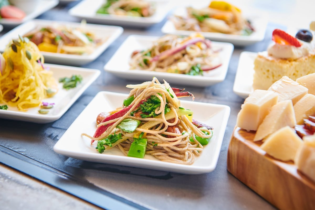 An assortment of dishes from the t4 escape lounge at Phoenix Airport, featuring a prominently a pasta dish with a cheese board and dessert cake on seperate platters to the right.