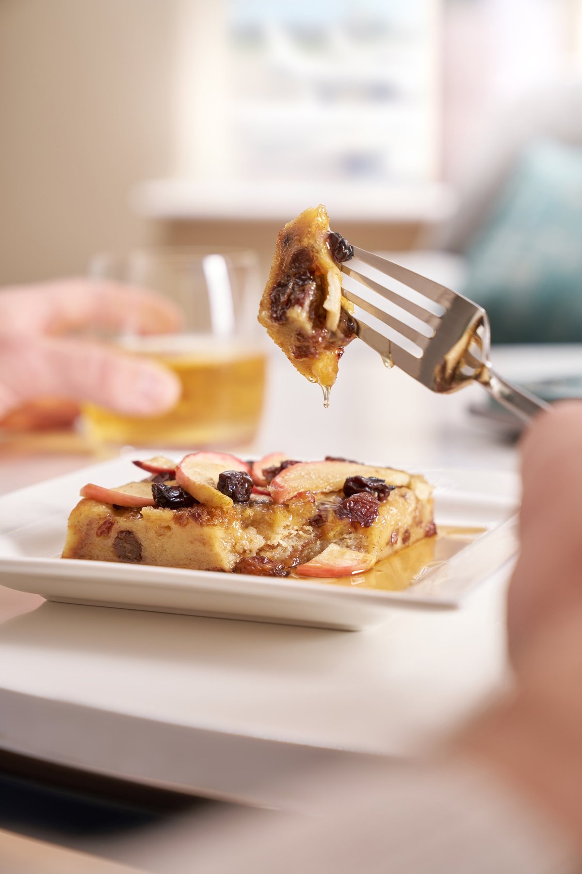 Close up image of a slice of bread pudding topped with apple slices and raisins, drizzled with syrup, on a white square plate. A fork is lifting a syrup-coated bite, with a blurred glass of juice or beer in the background.