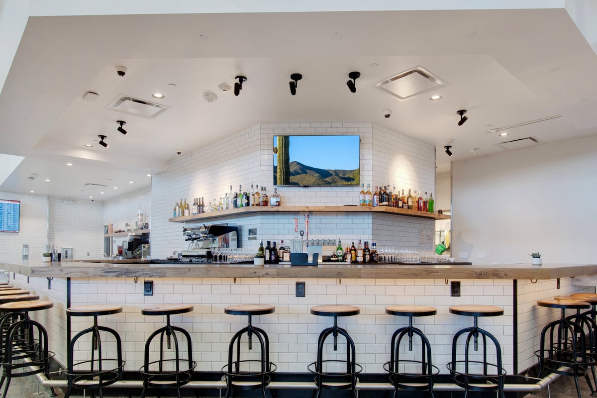 Bar area at the t3 escape lounge at phx airport, featuring a modern design with a white subway tile backdrop, wooden countertops and industrial style bar stools.