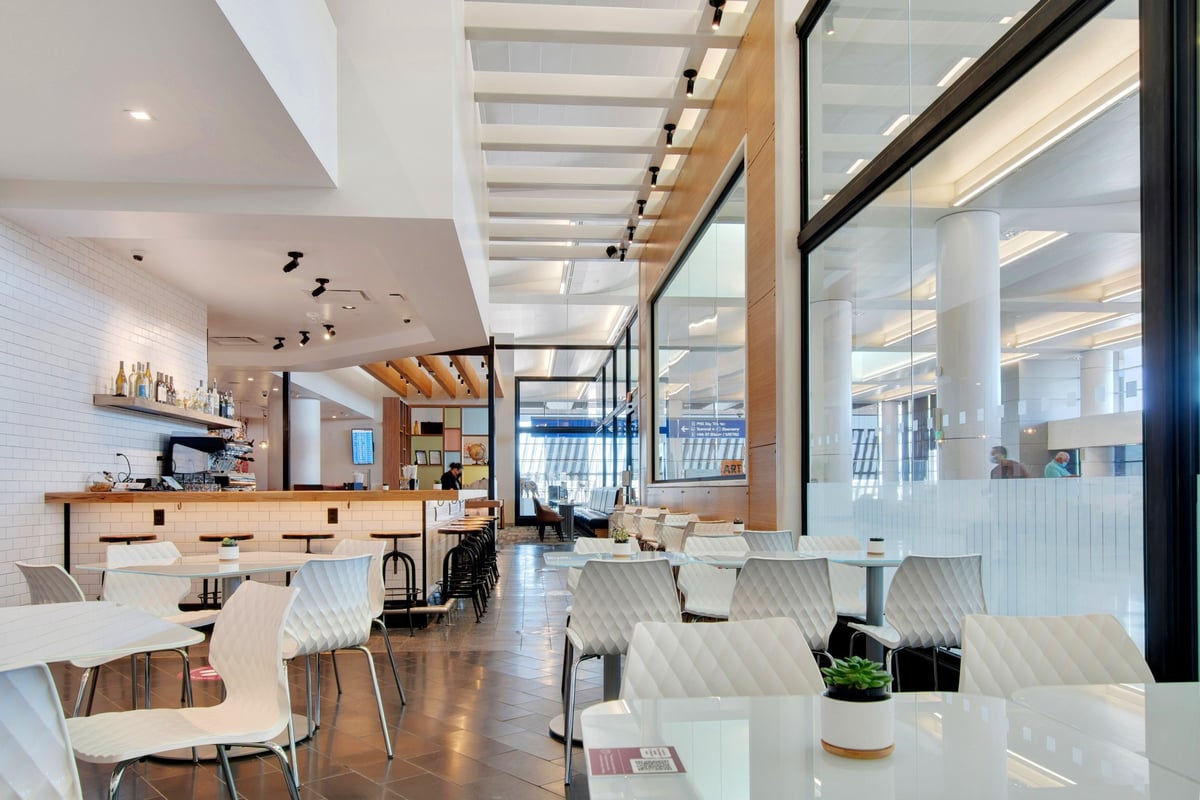 Dining area of the Escape Lounge at T3, PHX airport, featuring modern white chairs with quilted designs, sleek white tables, a bar counter with stools, and a coffee station.
