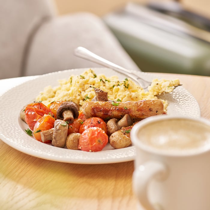 Food from the T3 Escape Lounge at PHX airport, featuring roasted tomatoes, mushrooms, sausages and scrambled eggs on a white plate with fork. A mug of coffee is pictured blurred in the foreground.