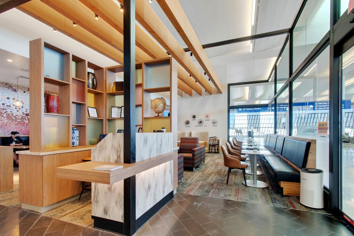Entrance and seating area of the escape lounge in T3 PHX airport. The space features modern wooden shelving with decorative items, a marble reception desk, seating with tables along glass windows and a bright open layout.