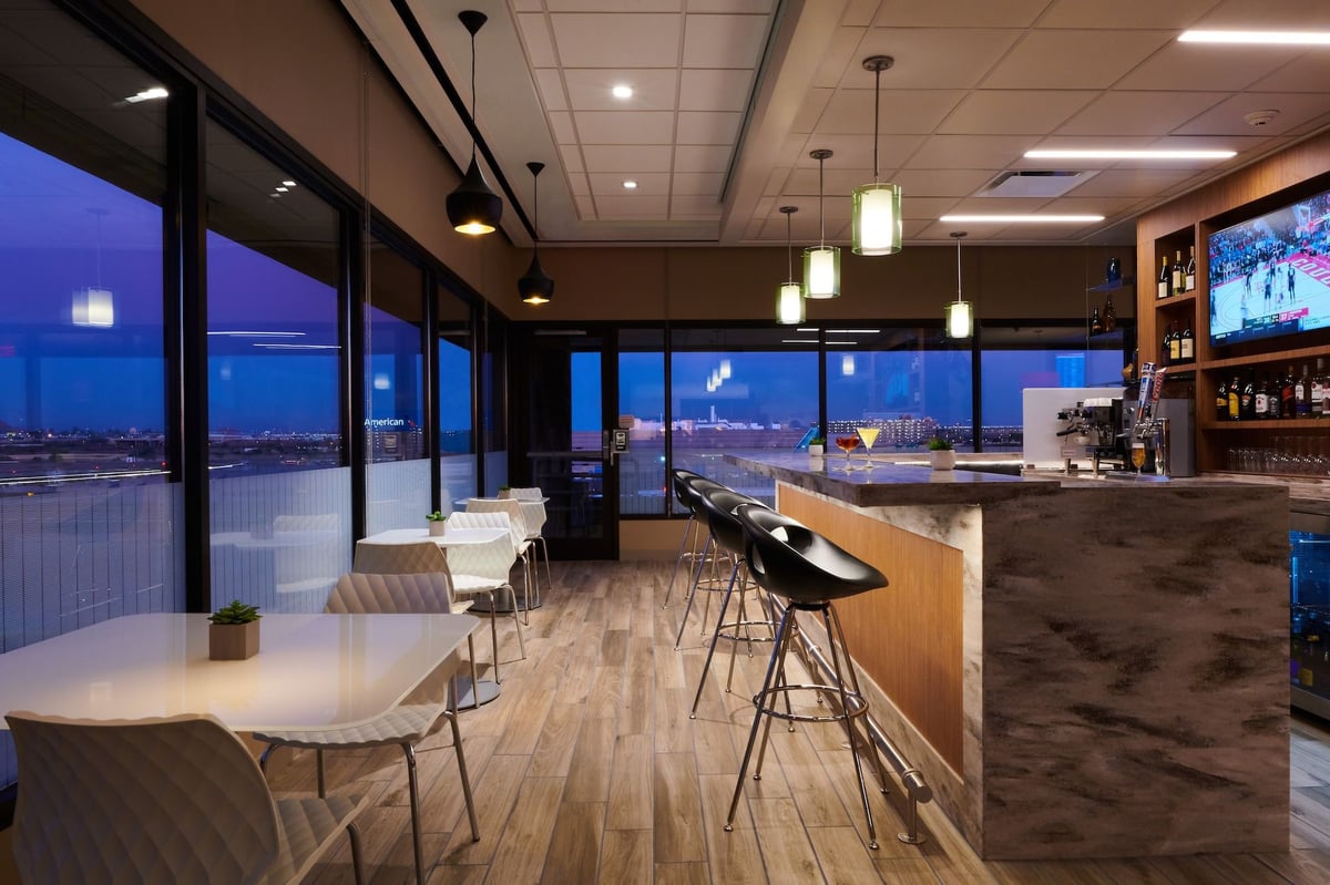 Image of bar and seating area at the T4 Escape Lounge at PHX Airport, featuring modern pendant lighting, sleek black barstools along a marble counter and white quilted chairs at dining tables along windows.