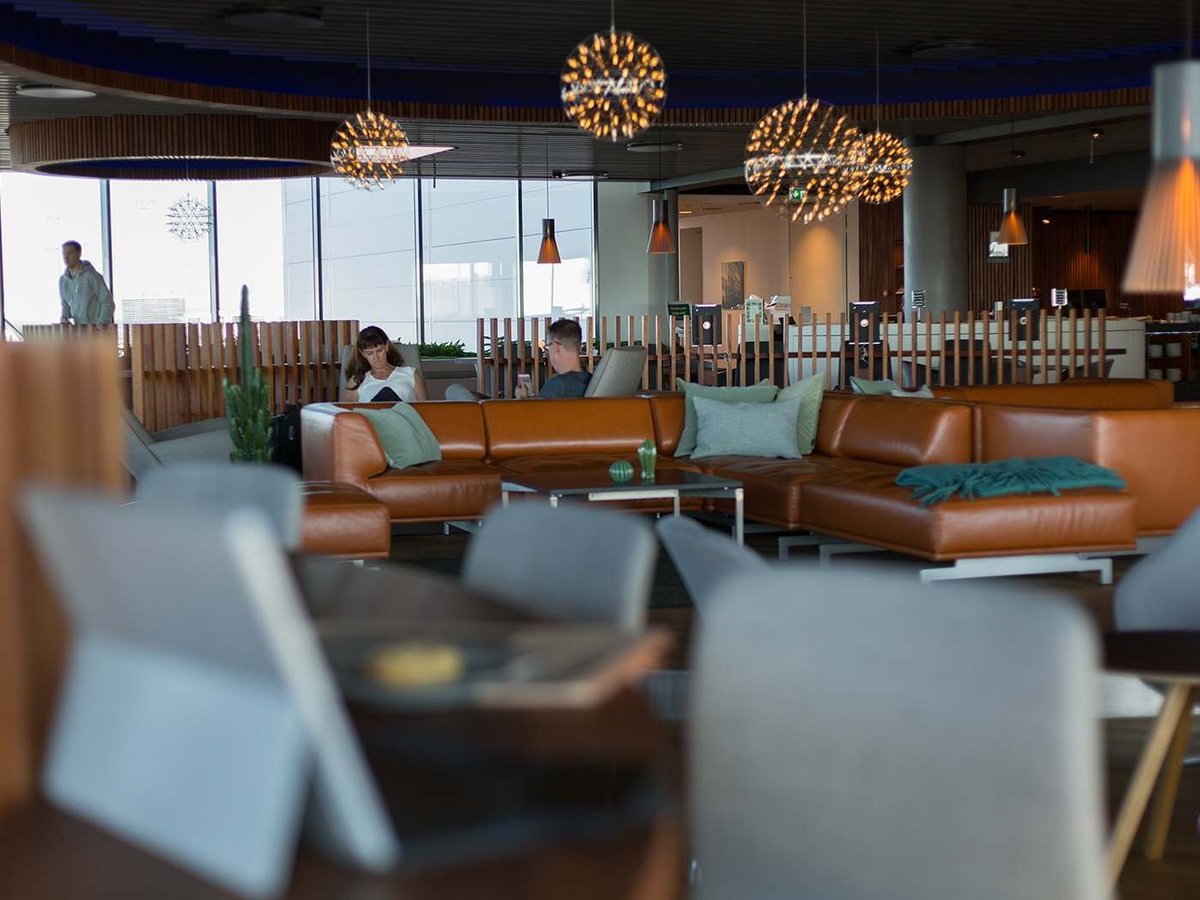 Interior view of the Eventyr Lounge at Copenhagen Airport, featuring spacious seating with luxurious tan leather sofas accented with teal cushions. The area is illuminated by unique spherical chandeliers, casting a warm, inviting glow.