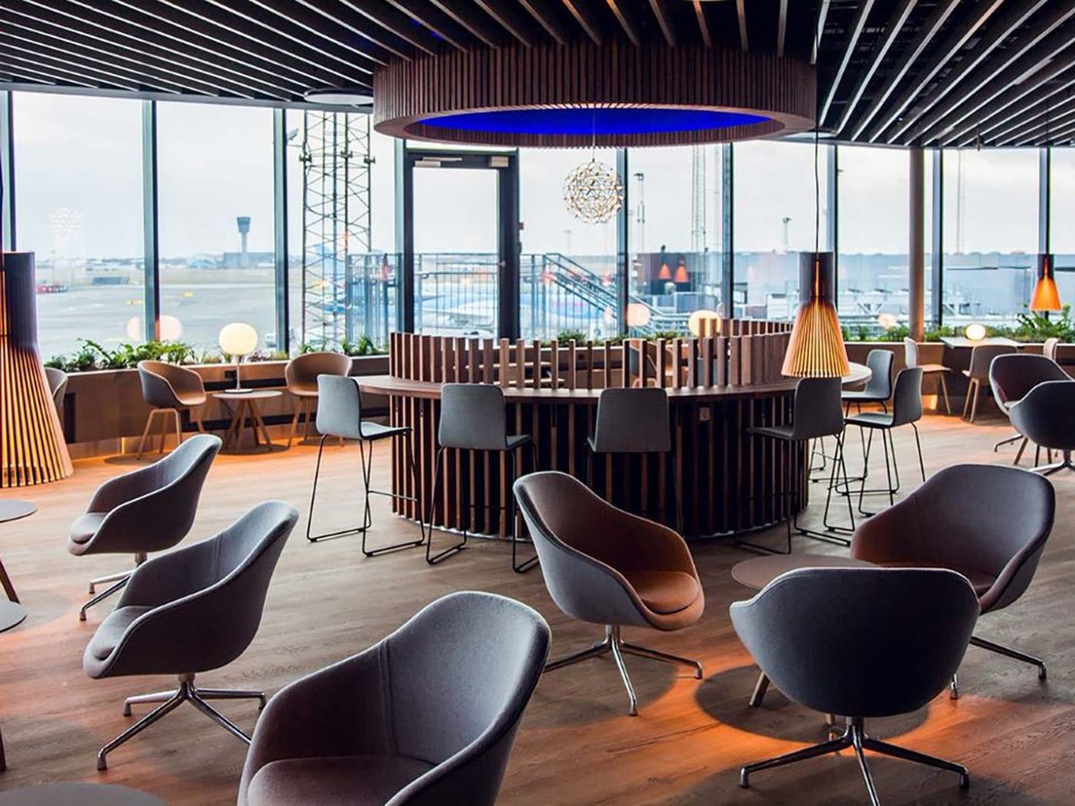 Spacious and elegantly designed Eventyr Lounge at Copenhagen Airport, featuring a variety of seating options including dark grey chairs and wooden tables. A striking blue LED light accents the bar area under a dark wooden ceiling, complemented by art