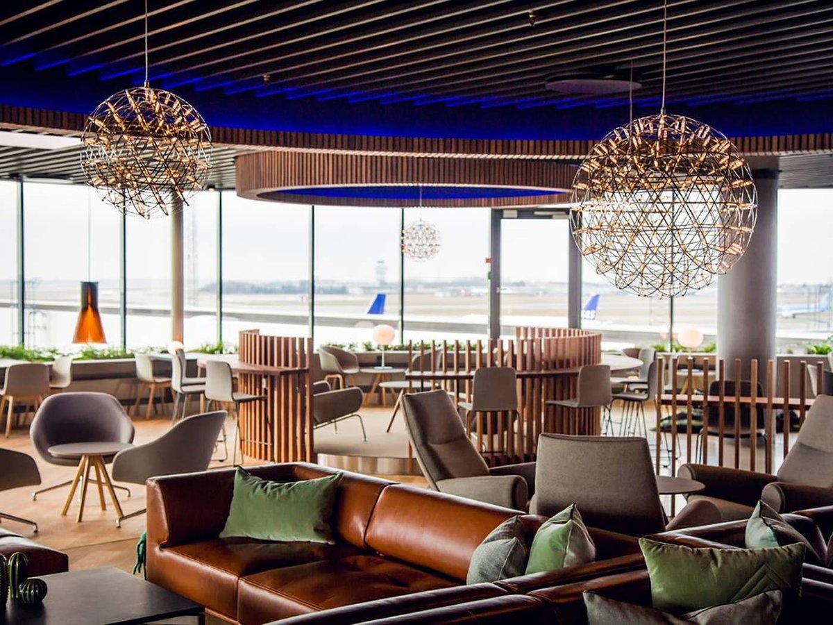 Interior of the Eventyr Lounge at Copenhagen Airport, showcasing a stylish and modern design. The spacious lounge features large spherical chandeliers made of intertwined metal wires hanging from a dark blue ceiling with wooden accents.