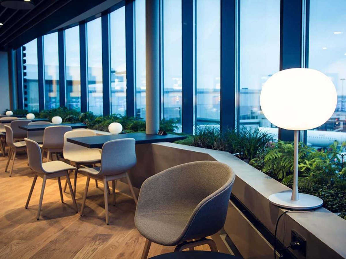 Modern seating area in Eventyr Lounge at Copenhagen Airport, featuring grey upholstered chairs along a curved countertop. A large globe lamp on a slender stand illuminates the space. Large windows provide a panoramic view of the airport runway.