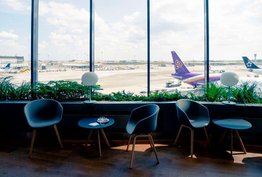 A bright and modern seating area inside the Eventyr Lounge, featuring sleek blue chairs and small round tables. The space is bathed in natural light streaming through large panoramic windows that offer a stunning view of the airport runway.