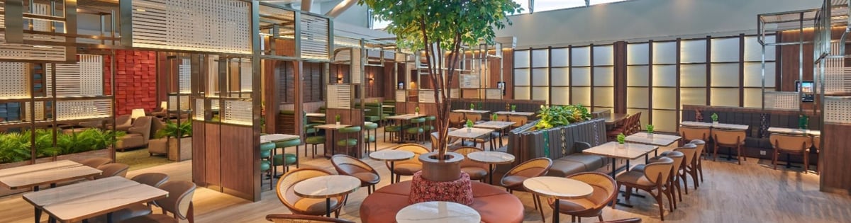 Seating area of the Plaza Premium Lounge in Kuala Lumpur Airport. The seating is pleasant browns, neutrals and greens, with some greenery in pots lining one side of a walkway, and some patchwork designs on wall dividers or wall decor.