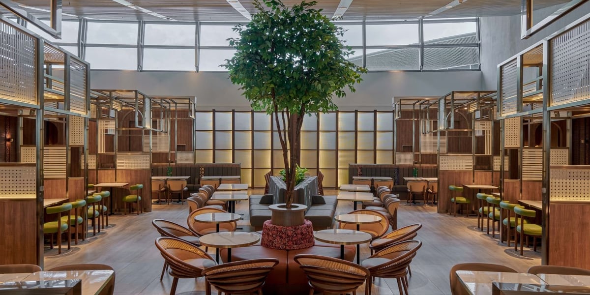 An open seating area at the Plaza Premium Lounge at Kuala Lumpur Airport. Large green foliage sits as a centrepiece to the room, surrounded by a variety of seating in brown, neutral and green colours.