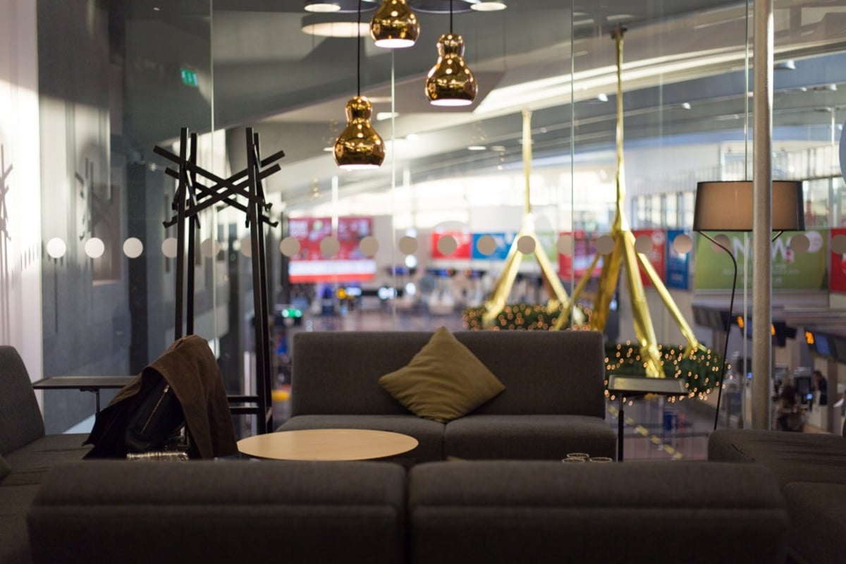 Image of dark grey sofas in the t2 pearl lounge at Arlanda Airport overlooking what appears to be the terminal. Gold light fixtures hang from the ceiling, illuminating the space. One of the sofas has an olive green cushion positioned in the middle.