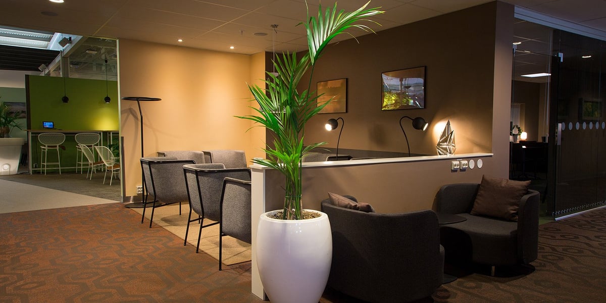 A cozy and modern seating area inside the Arlanda Airport t2 Pearl Lounge. The space features dark grey armchairs, soft lighting and a large potted plant in a sleek white vase. A partition wall separates different seating areas, with small desk lamps
