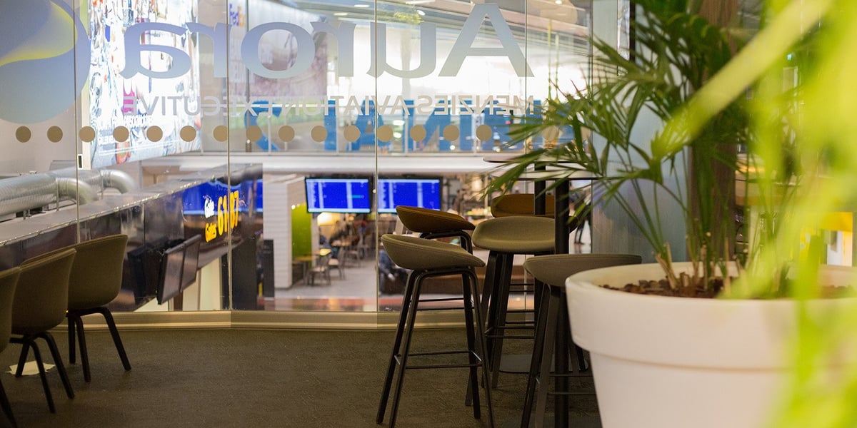 A seating area inside the Pearl Lounge at T2 Arlanda Airport, featuring modern high top stools and bar tables positioned next to a large glass wall overlooking the terminal. Lush green plants in pots add a natural element to the space.