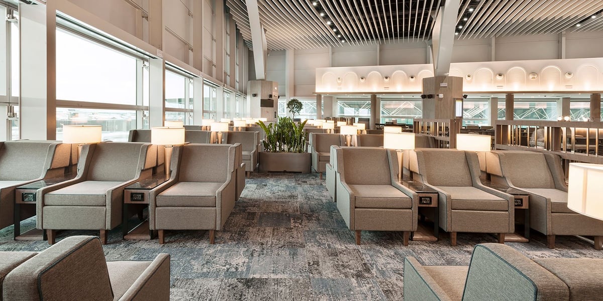 This image shows a spacious and elegantly designed seating area in the Plaza Premium Lounge at Fiumicino Airport. The lounge features rows of comfortable, boxy armchairs upholstered in a neutral gray fabric, each accompanied by a small lamp.