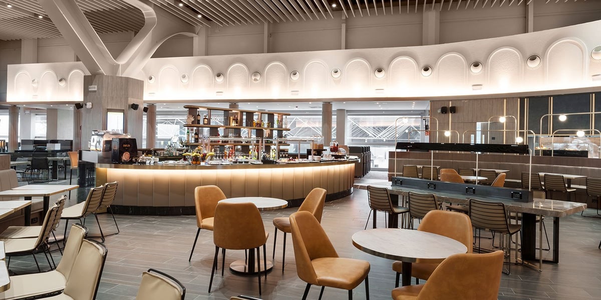 This image captures the sophisticated dining area within the Plaza Premium Lounge at Fiumicino Airport. The space features a contemporary design with a mix of high bar stools and plush, caramel-colored chairs grouped around stylish marble tables.
