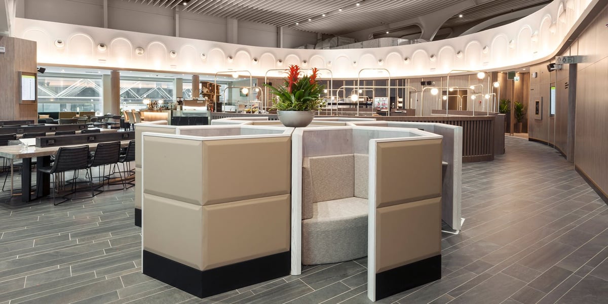 This image depicts another section of the Plaza Premium Lounge at Fiumicino Airport, focusing on the contemporary work and dining areas. At the forefront are modern workstations separated by high, beige privacy panels, providing guests with space.