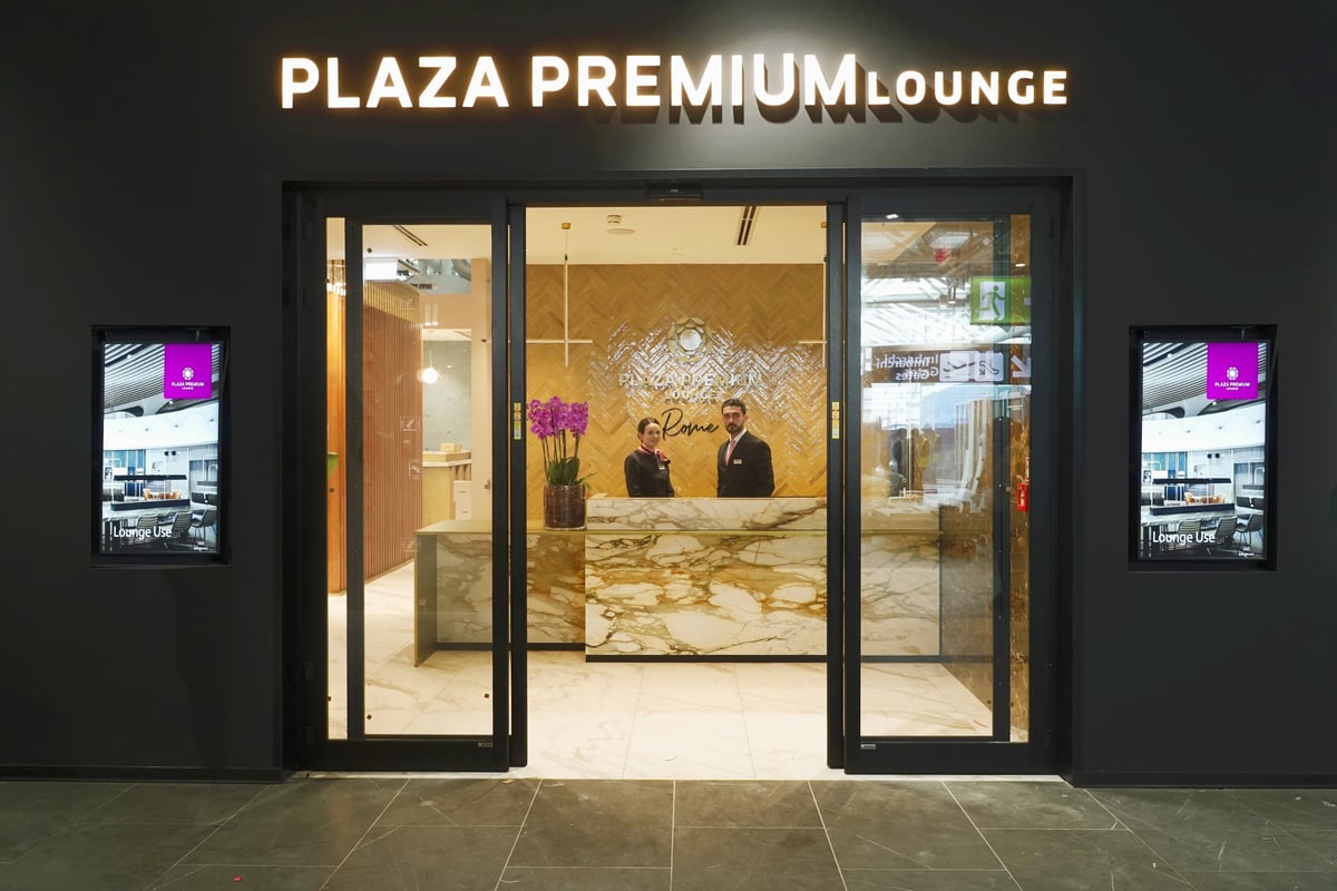 This image showcases the entrance to the Plaza Premium Lounge at Fiumicino Airport's Terminal 1. The entrance is marked by sleek glass doors framed by a dark facade, with the lounge's name illuminated in bright lettering above.