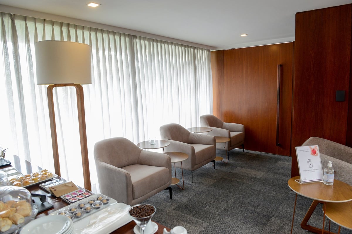 Buffet and seating area inside the landside Plaza Premium Lounge at São Paulo GRU Airport, featuring a selection of pastries and coffee, soft beige armchairs with side tables, floor-to-ceiling sheer curtains, and warm wood-paneled walls