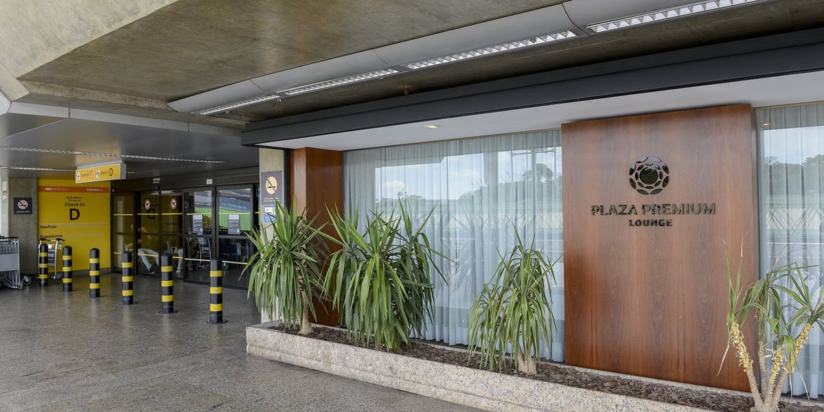 Exterior entrance of the Plaza Premium Lounge at São Paulo Guarulhos Airport (GRU), located landside near Terminal 2 Check-in D, featuring a sleek wood-paneled sign, large glass windows with sheer curtains, and tropical potted plants.