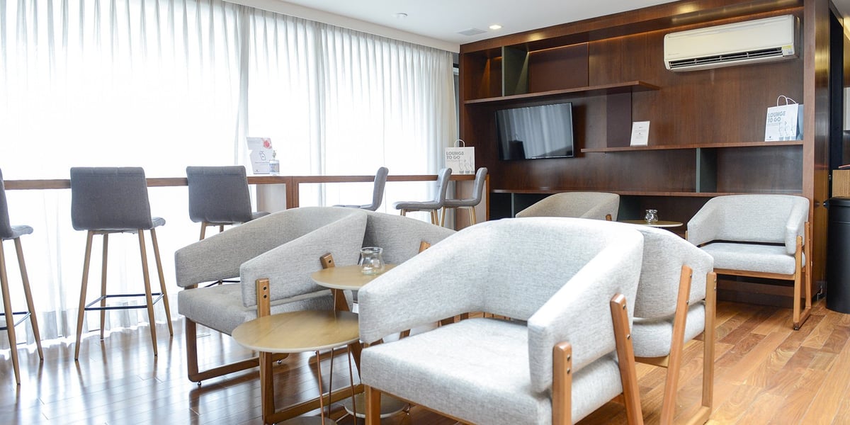 TV lounge area inside the landside Plaza Premium Lounge at São Paulo GRU Airport, featuring modern grey lounge chairs, high stools along a sunlit window counter, wooden flooring, built-in shelving, and a wall-mounted television.