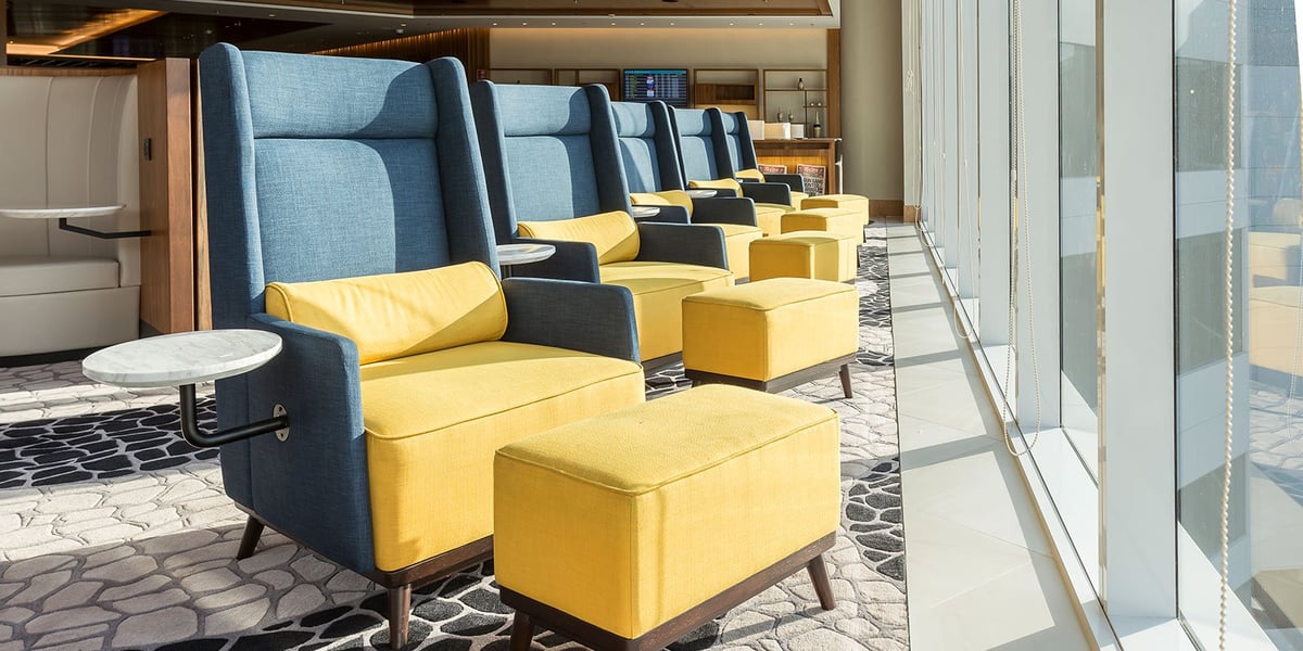 Sunlit relaxation zone at Plaza Premium Lounge in GIG Airport’s international terminal, featuring modern blue and yellow armchairs with footrests, individual marble side tables, and floor-to-ceiling windows overlooking the tarmac