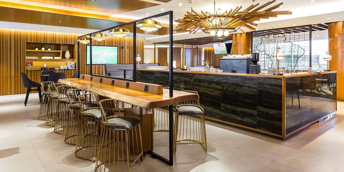 Modern bar and dining area inside Plaza Premium Lounge at Rio de Janeiro GIG International Airport, featuring a natural wood communal table with power outlets, gold-accented bar stools, designer lighting, and a stylish coffee and beverage station.