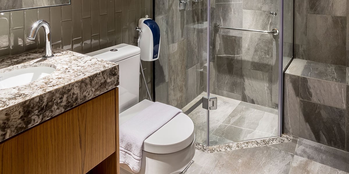 Modern private bathroom at the arrivals Plaza Premium Lounge in GIG Airport, featuring a glass-enclosed rainfall shower, granite countertop sink, bidet spray, and sleek tiled walls for a clean and upscale experience.