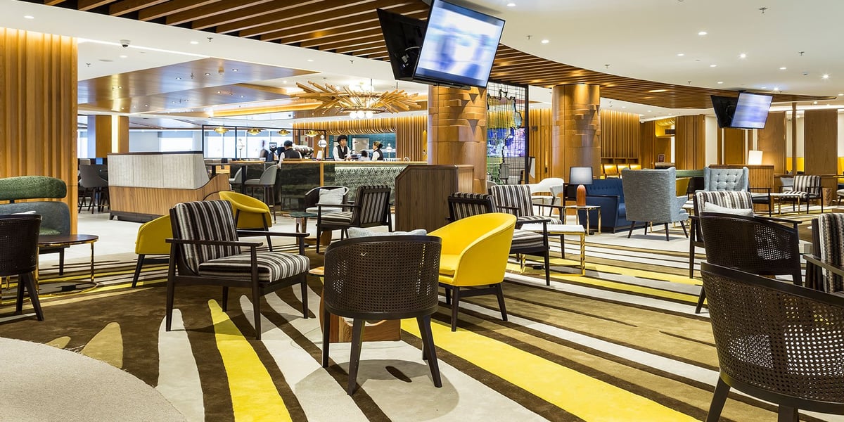 Vibrant dining and seating area at Plaza Premium Lounge in Rio de Janeiro GIG International Airport, showcasing a mix of modern armchairs, bold striped carpet, mounted TVs, and a well-lit service bar with contemporary wooden décor.
