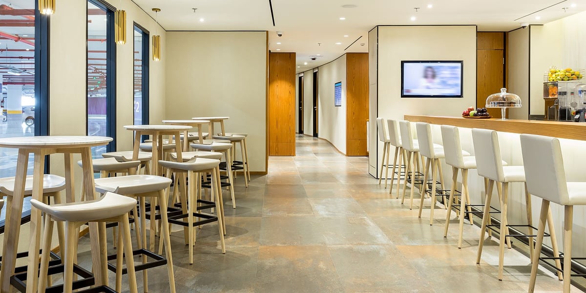Minimalist dining area at the arrivals Plaza Premium Lounge in GIG Airport, featuring high-top tables and bar seating with light wood finishes, a self-serve refreshment counter, and large windows overlooking the parking structure.