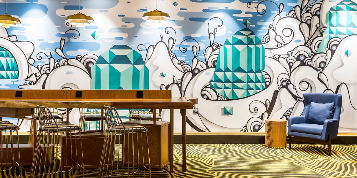 Colorful mural and contemporary seating area inside the domestic Plaza Premium Lounge at GIG Airport, featuring a striking gemstone-themed wall design, high-top communal table with charging ports, gold-accented stools, and a modern blue armchair.