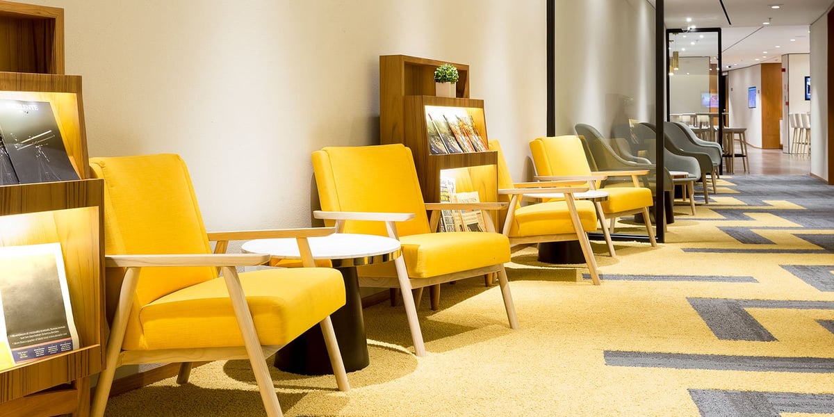Bright and inviting seating area at the arrivals Plaza Premium Lounge in Rio de Janeiro GIG Airport, featuring yellow armchairs with side tables, built-in magazine racks, and a patterned yellow and gray carpet in a calm, well-lit hallway.