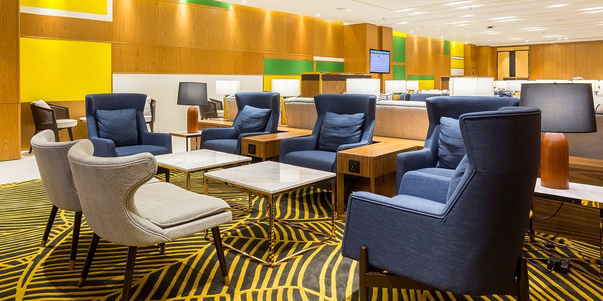 Comfortable seating area at the domestic Plaza Premium Lounge in Rio de Janeiro GIG Airport, featuring plush navy and beige armchairs, marble-top tables, wood panel walls with colorful accents, and ambient lighting for a relaxing atmosphere.