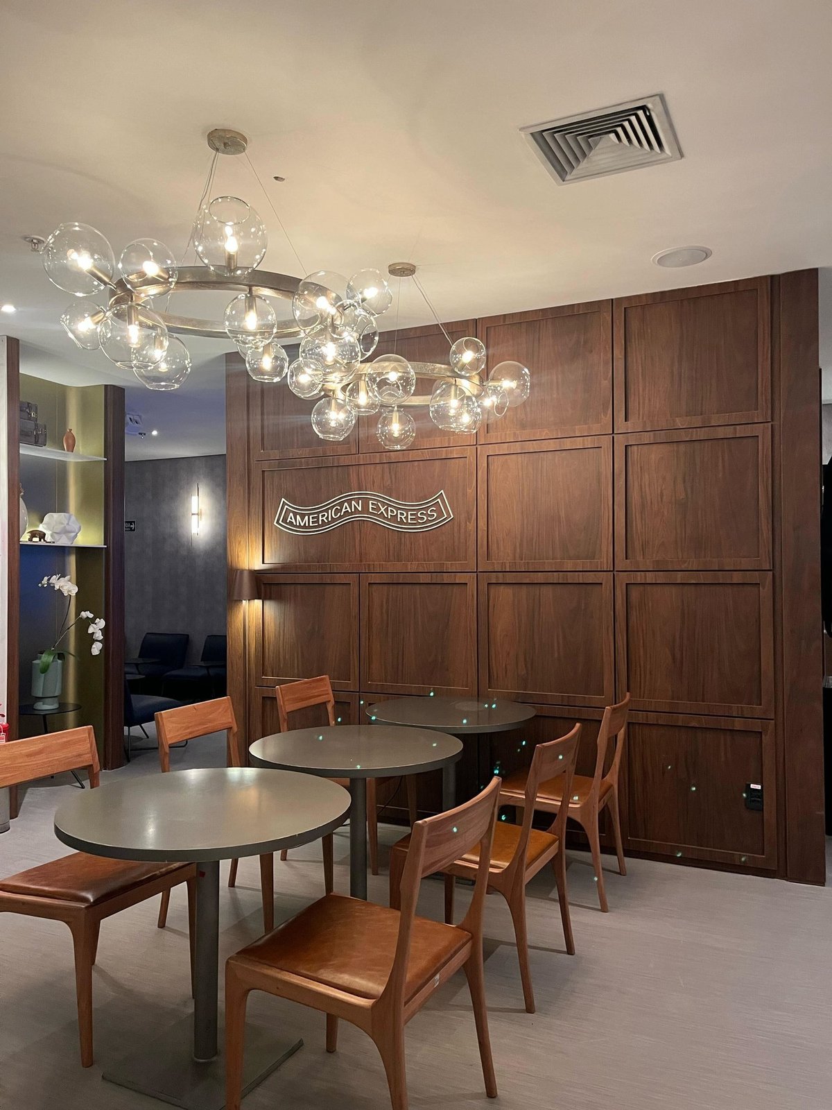 Dining area inside the 5th Avenue Lounge – W Premium at São Paulo GRU Airport, featuring round tables with wooden chairs, modern bubble chandeliers, and a warm wood-paneled wall with the American Express logo.