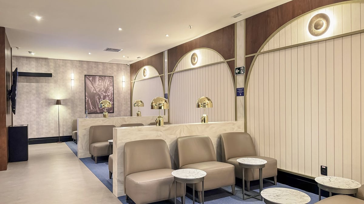 Sophisticated seating area at the 5th Avenue Lounge – W Premium in São Paulo GRU Airport, featuring beige leather chairs, marble-top side tables, golden dome lamps, and art-deco-inspired wall panels with warm lighting.