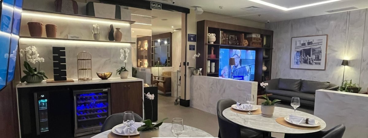 Dining and lounge area at the 5th Avenue Lounge – W Premium in São Paulo GRU Airport, featuring marble-top tables set with glassware, elegant shelving with decorative accents, a wine cooler, leather sofas, and a wall-mounted TV.