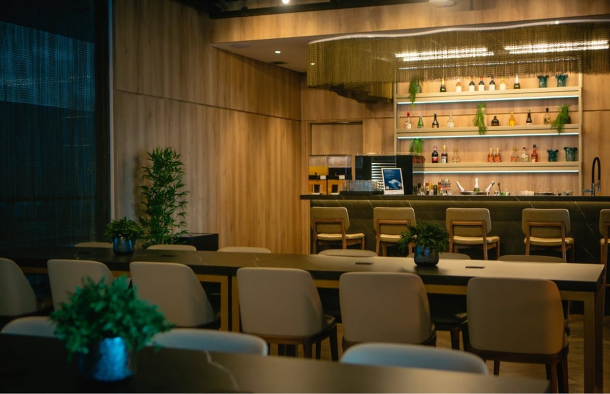 Sophisticated bar area at The Pier – W Lounge in Terminal 3 of São Paulo GRU Airport, featuring a modern wooden bar counter with high stools, illuminated shelves stocked with premium spirits, potted greenery, and sleek dining tables for guests.