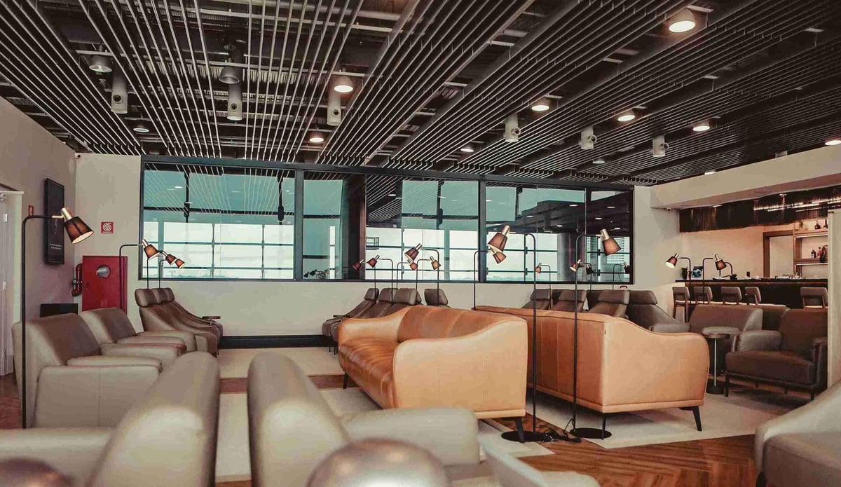 Seating area at The Pier – W Premium Lounge in Terminal 3 of São Paulo GRU Airport, featuring tan and gray leather armchairs with personal reading lamps, modern ceiling lighting, and a view of the bar area in a spacious, contemporary setting.