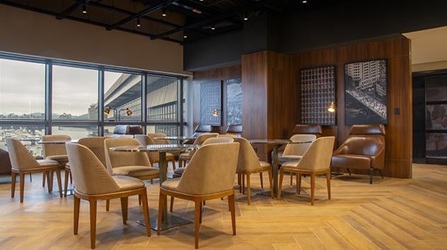 Dining area at W Premium Lounge – The West, Terminal 2 of São Paulo GRU Airport, featuring elegant leather chairs, wooden tables, parquet flooring, floor-to-ceiling windows with apron views, and modern wall art for a refined ambiance.