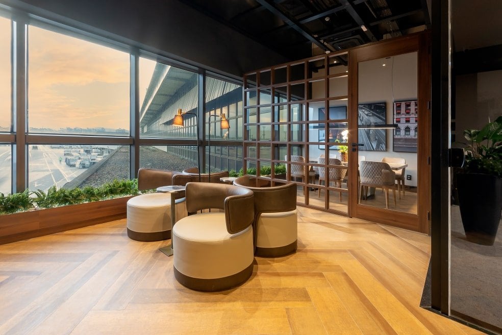 Modern seating area at W Premium Lounge – The West, Terminal 2 of São Paulo GRU Airport, featuring round leather chairs, wood flooring, floor-to-ceiling windows with tarmac views, and a partitioned meeting space in the background.