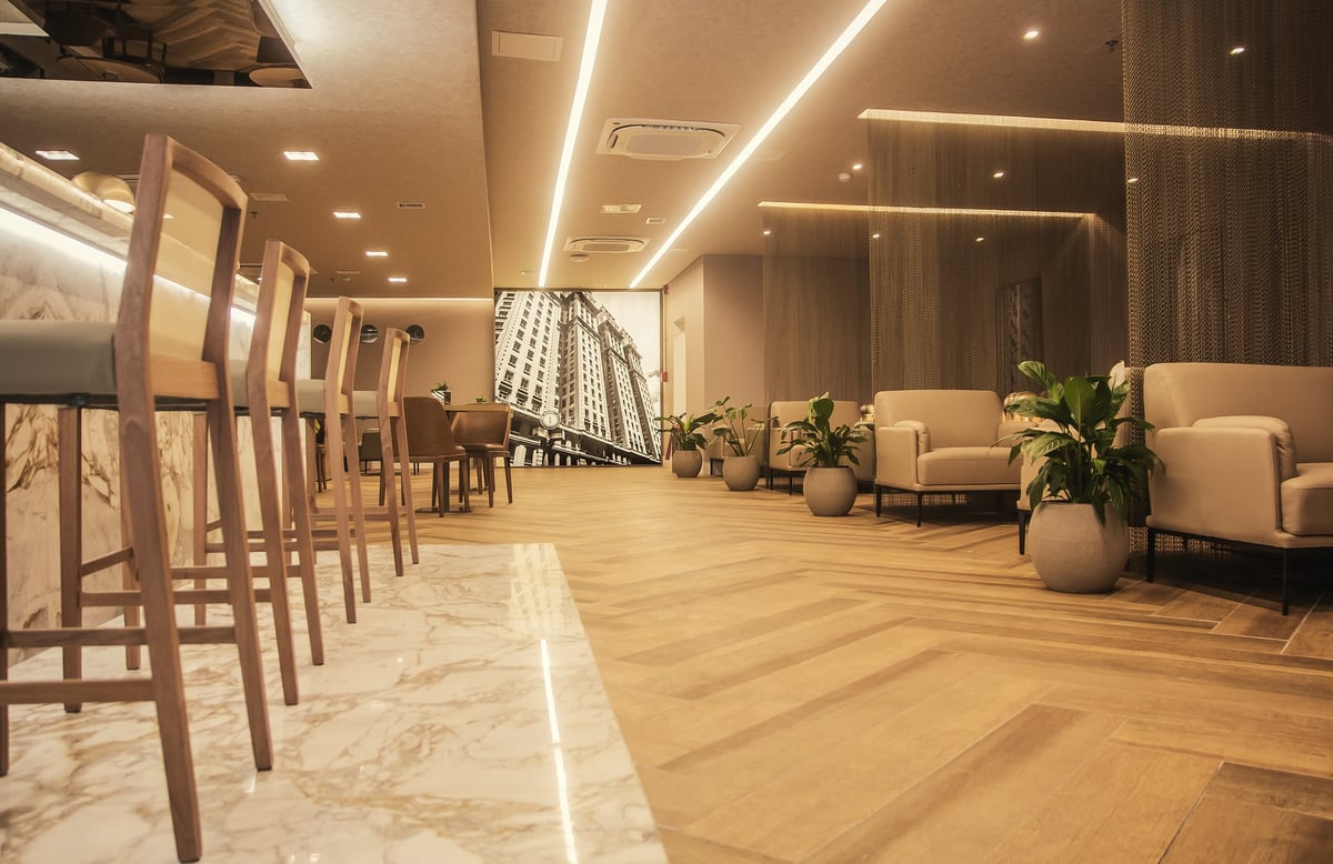 Spacious interior of W Premium Lounge at Terminal 1 of São Paulo GRU Airport, showcasing a marble bar with high stools, modern lounge seating with neutral-toned armchairs, decorative potted plants, warm wood flooring, and a large mural