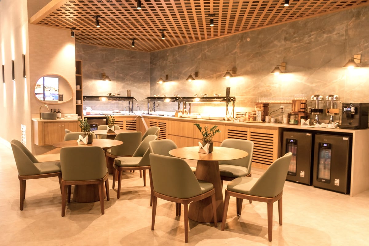 Buffet area at W Premium Lounge in Porto Alegre Airport featuring a modern dining setup with round wooden tables, green upholstered chairs, ambient lighting, and a self-service food and beverage counter with hot dishes, drinks, and coffee stations.